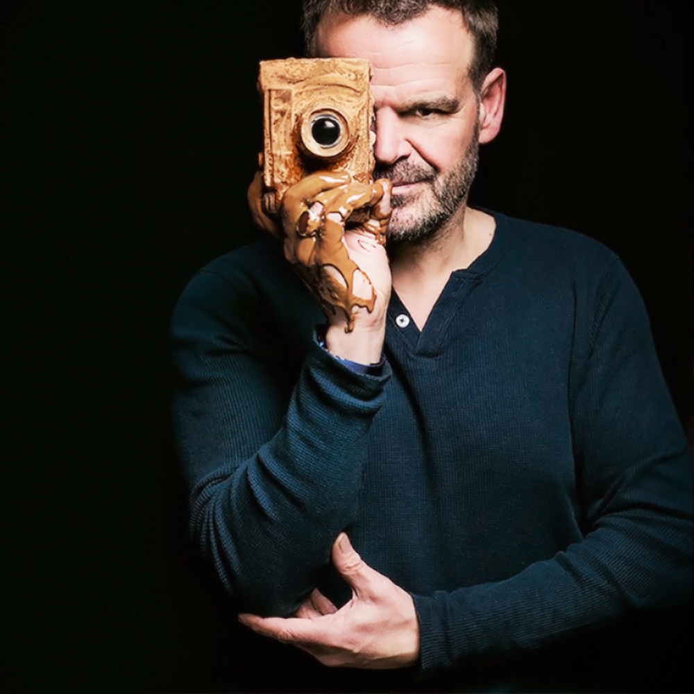 George Zarzonis poses, holding a camera, covered in chocolate